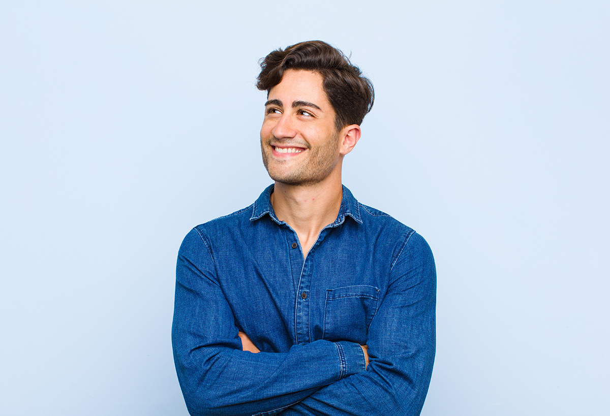 young handsome man feeling happy, proud and hopeful, wondering or thinking, looking up to copy space with crossed arms against blue background
