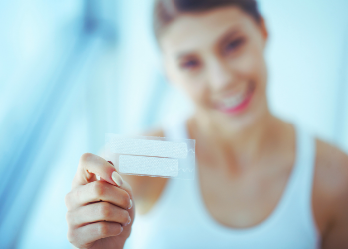 Person in white tank top holding a transparent adhesive bandage strip between thumb and index finger; blurred background emphasizes the bandage. Let me know if you’d like a version adapted for first aid education, product packaging, or pediatric care.